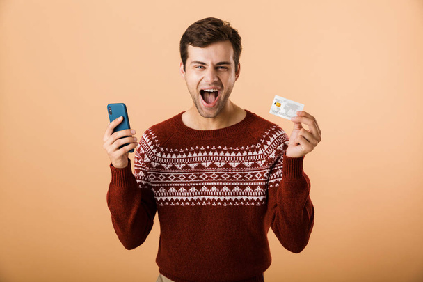 Portrait a joyful young man dressed in sweater standing isolated over beige background, holding mobile phone, showing credit card - Fotó, kép