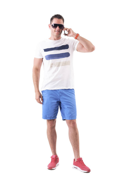 Happy man in shorts and t-shirt relaxing and talking on cellphone. Full body isolated on white background.  - Fotó, kép