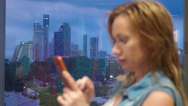мокра блондинка стоїть біля вікна на фоні хмарочосів, використовує свій телефон, поки йде дощ за вікном
 - Кадри, відео