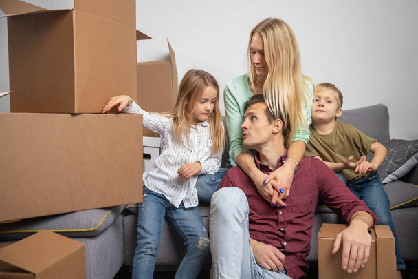 Photo of happy man, woman, girl and boy sitting on gray sofa among cardboard boxes for moving - Photo, Image