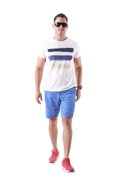 Fresco jovem adulto feliz em t-shirt e shorts de verão com óculos de sol andando e sorrindo. Corpo inteiro isolado em fundo branco
.  - Foto, Imagem