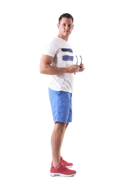 Vista lateral do jovem vestindo camiseta e tiros segurando óculos de sol olhando para a câmera. Corpo inteiro isolado em fundo branco
.  - Foto, Imagem