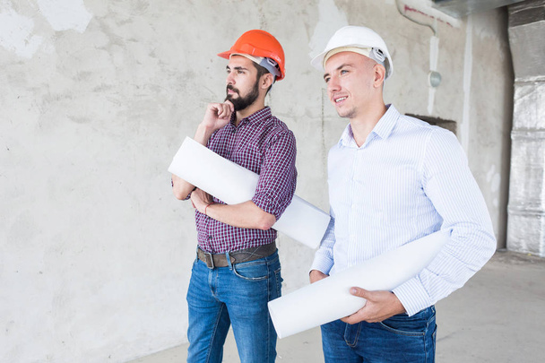 male engineers, architects in helmets stand by the window and hold a roll of paper with a building drawing - Foto, afbeelding