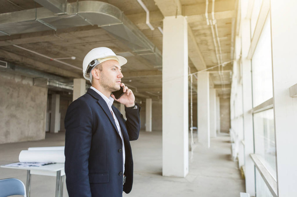 male engineer, foreman in a helmet, in a business suit with paper with drawings, in the construction being completed talking on mobile phone. building supervision - Foto, afbeelding
