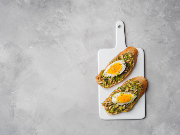 Whole-grain toasts plated avocado with eggs and fresh mini herbs with sunflower seeds and sprinkled black salt served on ceramic white board over grey concrete background, top view, Diet food concept, gluten-free  - 写真・画像
