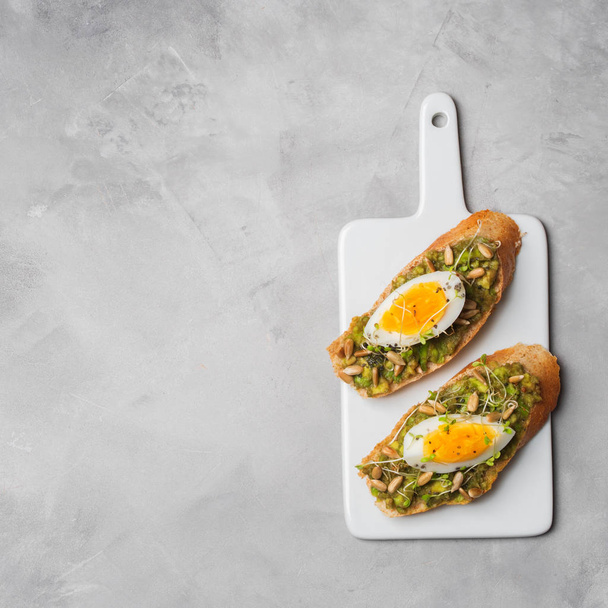 Whole-grain toasts plated avocado with eggs and fresh mini herbs served on ceramic white board over grey concrete background, top view, Diet food concept, gluten-free  - 写真・画像