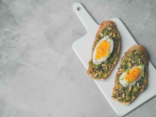 Whole-grain toasts plated avocado with eggs and fresh mini herbs with sunflower seeds served on ceramic white board over grey concrete background, top view, Diet food concept, gluten-free  - Foto, imagen