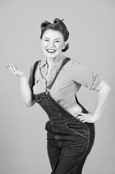 Portrait of smiled and cheerful pin-up girl standing with arm on hips and holding copy space on her palm isolated over gray background. Brunette happy woman in denim with open palm hand. - Φωτογραφία, εικόνα