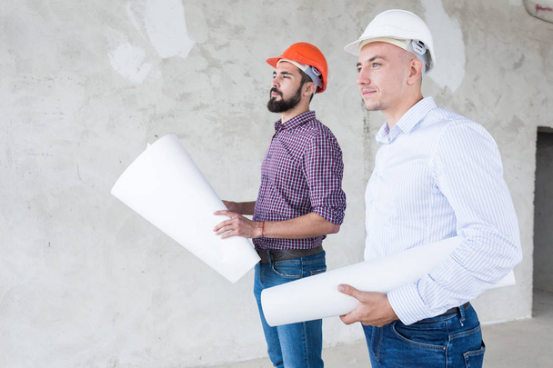 male engineers, architects in helmets stand by the window and hold a roll of paper with a building drawing - Foto, Imagem