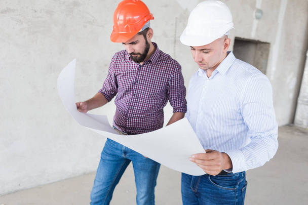 male engineers, architects in helmets stand by the window and hold a roll of paper with a building drawing - Фото, изображение