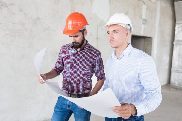 male engineers, architects in helmets stand by the window and hold a roll of paper with a building drawing - Foto, Imagen