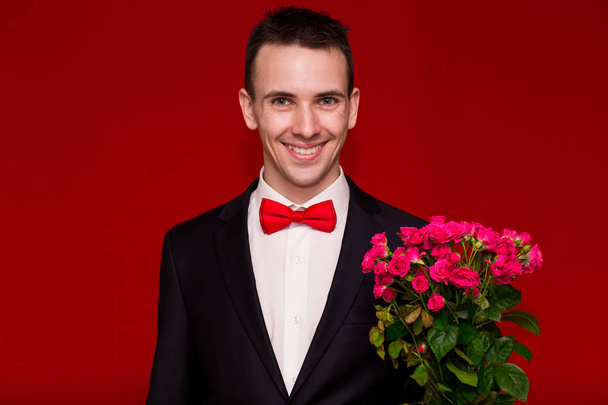 Καλαίσθητο άνθρωπος στο κοστούμι κρατώντας ένα μπουκέτο λουλούδια με κόκκινο φόντο - Φωτογραφία, εικόνα