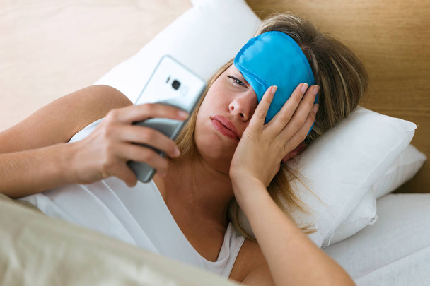 Shot of tired young woman lying in bed pulling up sleeping mask to see what time is it at smartphone in bedroom at home. - Photo, image