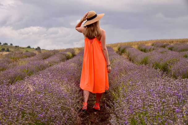 A girl in a bright orange dress and straw stands in a field of lavender at dawn with her back to the photographer sunset - 写真・画像