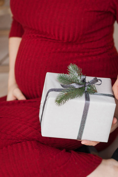 A pregnant woman in a red dress holds in her hands a box with a New Year's gift. Close up - Фото, изображение