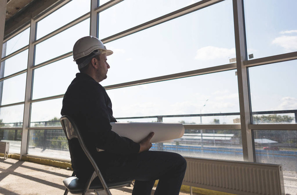 the builder architect in a helmet with drawings is working at the construction site. supervision of construction. silhouette by the window - Zdjęcie, obraz