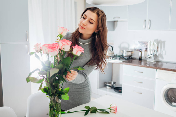 Vrouw zet roze rozen in vaas. Jonge huisvrouw verzorgen van gezelligheid in de keuken. Moderne keukenontwerp. Wit en zilver keuken - Foto, afbeelding