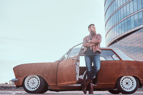Stylowe Brodaty mężczyzna w staromodny ubrania, opierając się na dostrojony samochód retro stary z otwartych drzwi w mieście parkowanie w pobliżu wieżowca. - Zdjęcie, obraz