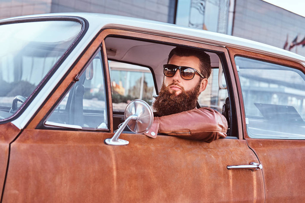 Varón barbudo en gafas de sol vestido con chaqueta de cuero marrón que conduce un coche retro
. - Foto, Imagen