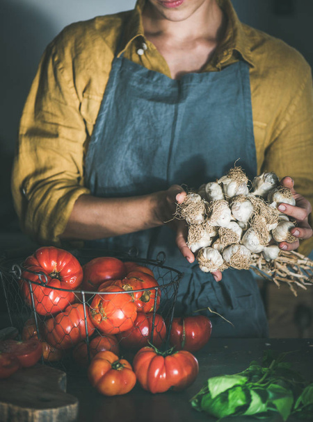 Woman in linen apron holding bunch of garlic for cooking tomato sauce, canned tomatoes or pasta with basil and heirloom tomatoes at kitchen counter. Healthy cooking, slow food or comfort food - Fotografie, Obrázek