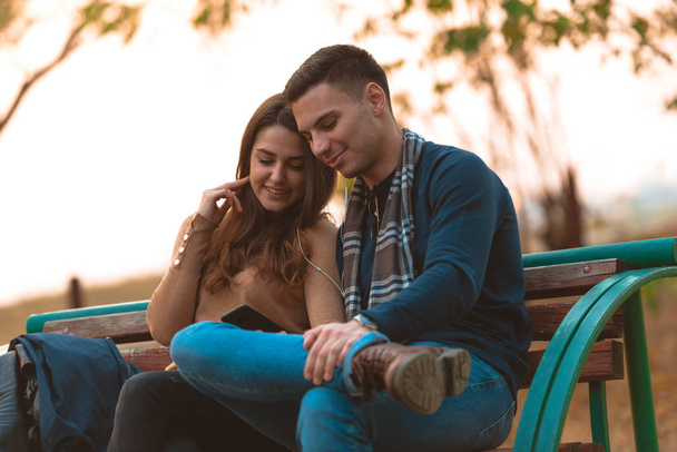 Young couple listening to music,smiling,looking at the phone and sitting on a bench in park. - Photo, Image