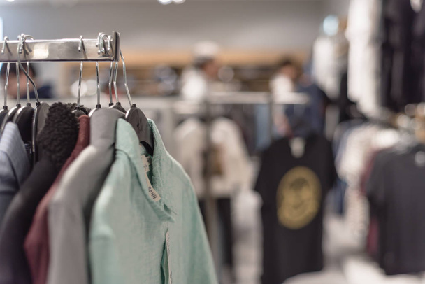Selective focus, shallow dof image of clothes shop with trendy clothing on hanger and blurry customer shopping, selecting textile product in background. - Zdjęcie, obraz