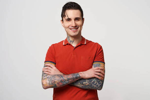 Ένας μελαχροινός νεαρός με τα μαλλιά του διπλωμένα και ένα ευτυχισμένο χαμόγελο, φορώντας ένα κόκκινο πόλο T-shirt στέκεται με τα χέρια σταυρωμένα, έχει ένα τατουάζ στο λαιμό, χέρια και πάνω από το λευκό φόντο - Φωτογραφία, εικόνα