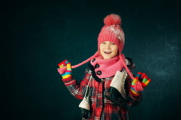 Studioporträt eines kleinen Mädchens in Jacke und Strickmütze auf dunklem Hintergrund. Kind mit Schlittschuhen - Foto, Bild
