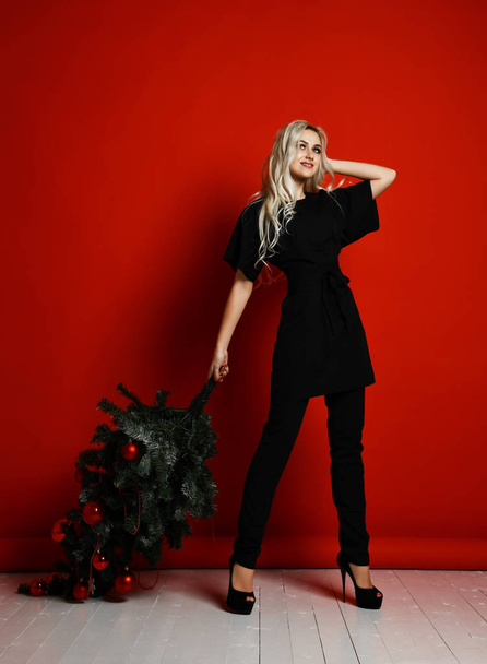 Blonde woman in black cloth hold xmas Christmas tree on her shoulders - Zdjęcie, obraz