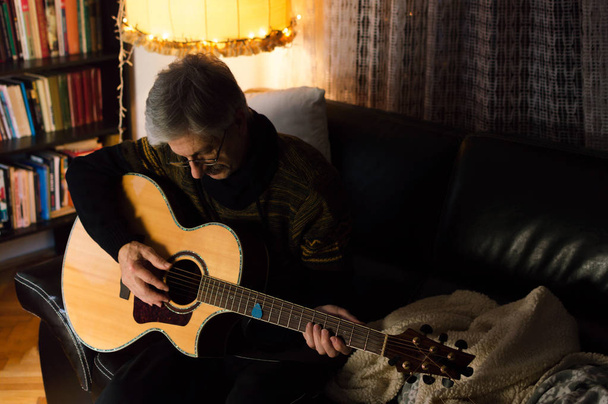 Γέρος παίζει ακουστική κιθάρα στο καναπέ του σαλονιού - Φωτογραφία, εικόνα