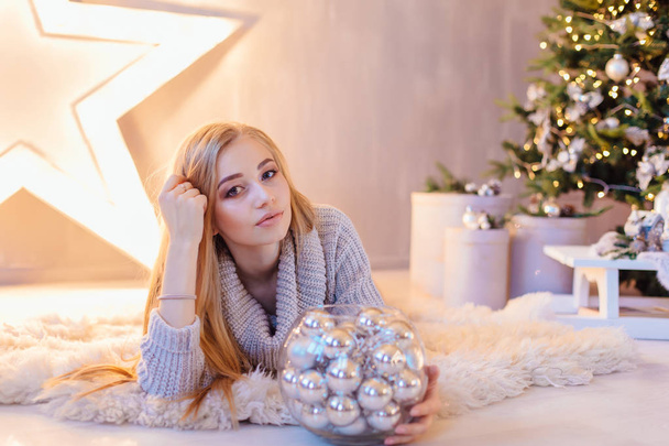 Beautiful young blond woman dressed in white sweater laying next to the big star with bowl full of silver balls in a holiday interior with Christmas tree. - Photo, Image