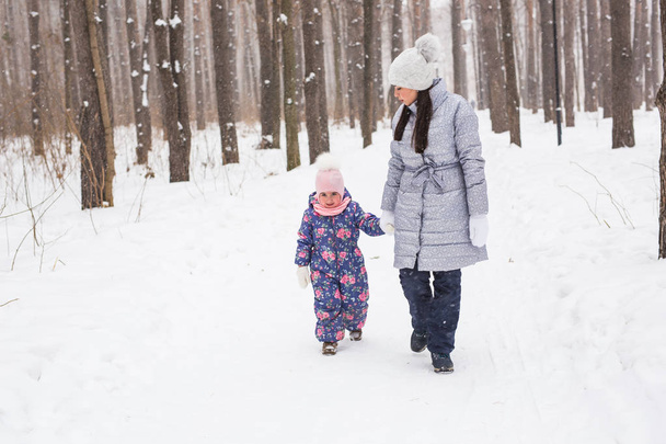 Family, winter and nature concept - Young cheerful mother with cute little daughter walking in the snow park - Photo, Image