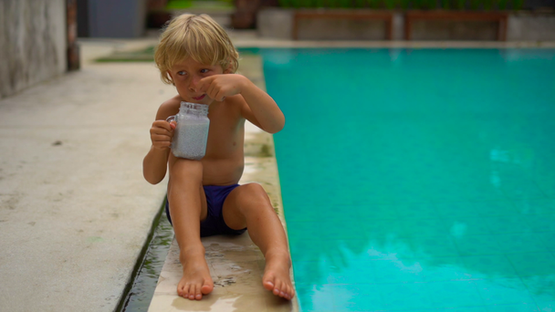 Slowmotion βολή του το μικρό αγόρι τρώει μια πουτίγκα chia κάθεται σε μια πλευρά της πισίνας - Πλάνα, βίντεο