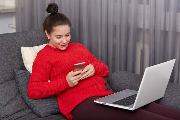 Pregnant woman has dark hair combed in bun, types text message on cellular, surf social networks, sits at couch with modern electronic gadgets, feels relaxation. Motherhood and technology concept - Fotografie, Obrázek