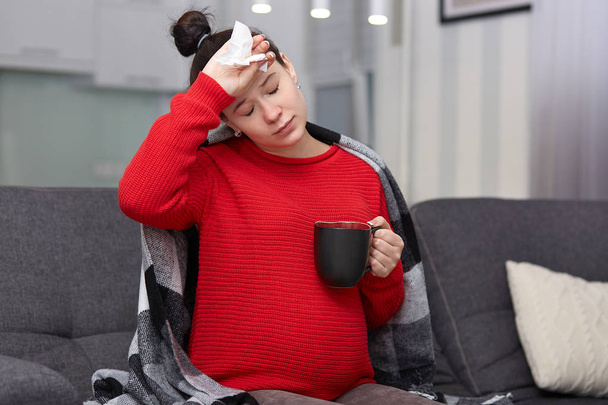 Photo of stressful woman has sick expression, keeps hand on forehead as suffers from headache, holds tissue and mug of tea with honey, cures disease at home, being pregnant. Illness and health care - Φωτογραφία, εικόνα