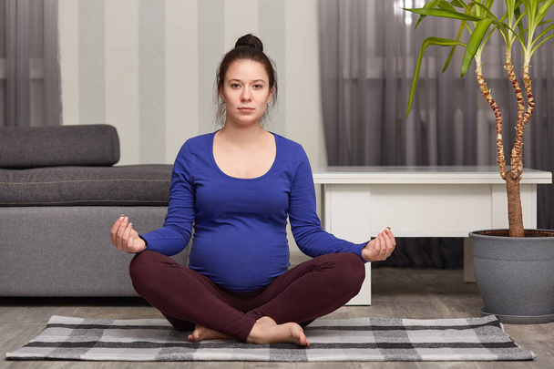 Photo of attractive dark haired expectant mother sits in lotus pose, practices yoga at home, wears comfortable clothes, sits on floor in modern apartment, meditates indoor, concentrates. Zen posture - Valokuva, kuva