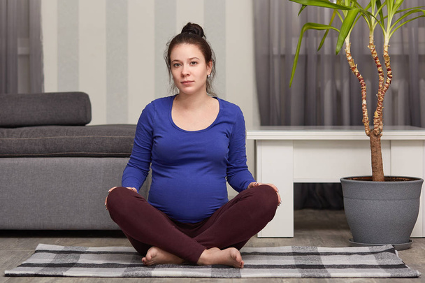 Indoor shot of beautiful European pregnant woman sits in zen posture, keeps legs crossed, has yoga exercises at home, looks directly at camera. People, pregnancy, sport and wellness concept. - Photo, image