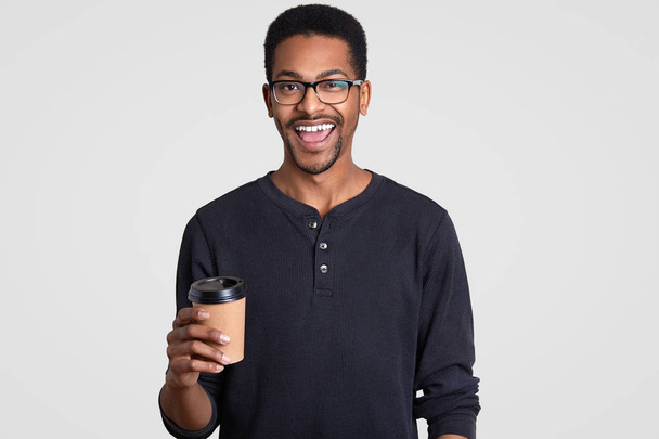 Image of happy smiling teenge boy with dark skin, wears spectacles, holds takeout coffee, enjoys fresh beverage, dressed in casual jumper, models against white background. People and coffee break - Φωτογραφία, εικόνα