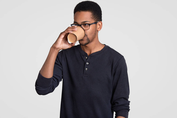Serious prosperous businessman has coffee break, holds paper cup, enjoys hot drink, wears glasses and casual jumper, stands indoor against white background. People, ethnicity and drinking concept. - Foto, imagen