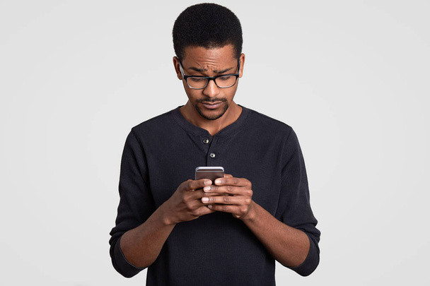 Condused black Afro African man downloads file on cellular, looks with puzzlement in screen, wears optical glasses, dressed in casual clothes, isolated over white background. Guy with cell phone - Photo, Image