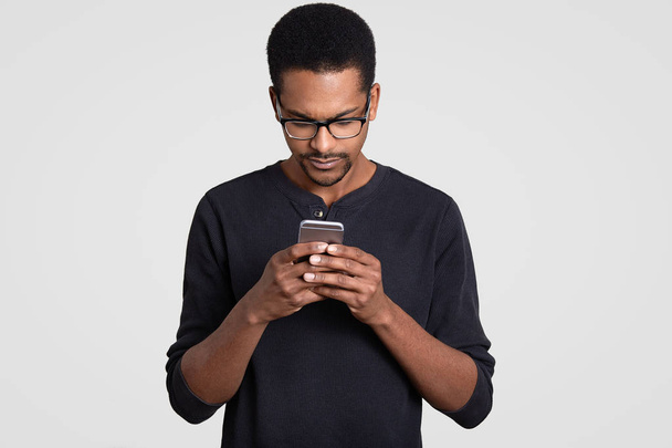 Photo of concentrated dark skinned handsome guy reads news in internet, holds cell phone, wears transparent glasses and sweater, isolated over white background, checks email box, uses free internet - Fotografie, Obrázek