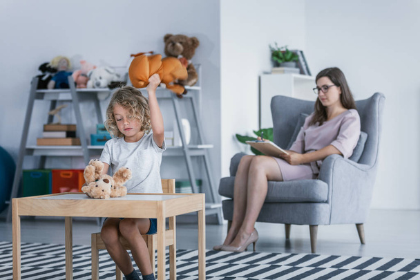 A child with behavioral problems sitting by a table and hitting a teddy bear during a therapeutic meeting with a therapist in a family support center. - Foto, Imagem