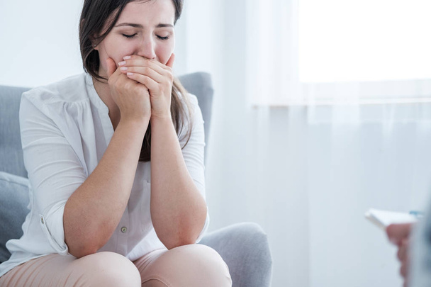 A stressed crying young woman covering her mouth with hands while sitting in a bright room. Empty space in the background. - Photo, image