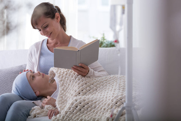 Lächelnde Mutter liest einem krebskranken Kind mit Kopftuch Buch vor - Foto, Bild