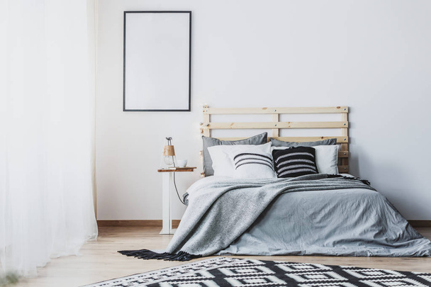 Mockup of empty poster in white bedroom interior with grey wooden bed with cushions. Real photo - Фото, зображення