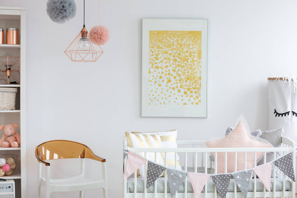 Stylish white and brown chair next to crib with pillows in trendy baby room with golden poster on the wall - Фото, изображение