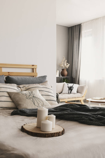 Blurred foreground with candles on wooden trunk tray in real photo of bright open space bedroom interior with window with drapes and couch - Фото, зображення