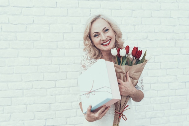 Woman in White Dress with Gift Box and Bouquet. Young Woman. Long Hair. Standing Woman. Red Tulip. White Flower. Celebration Concept. Holiday in March. Holding Bouquet in Hand. Brick Wall. - Foto, immagini