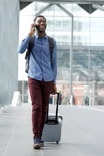 Full body portrait of young black man traveling with bags and talking on phone at station - Photo, Image