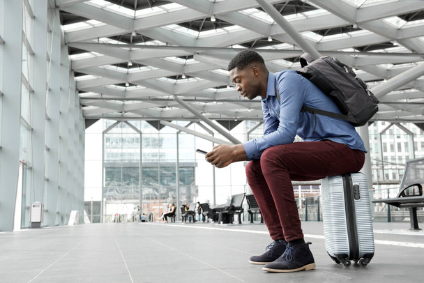 Retrato lateral del hombre afroamericano sentado en la maleta en la estación con teléfono móvil
 - Foto, Imagen